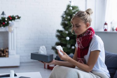Sıcak eşarplı hasta bir kadın arka planda Noel ağacının yanında kağıt peçete tutuyor.