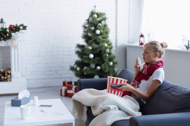 TV kumandalı hasta bir kadın ve bulanık noel ağacının yanındaki kanepede öksüren patlamış mısır kovası.