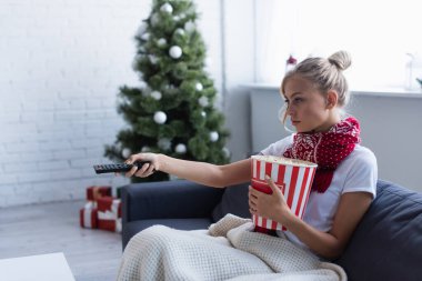 ill woman with bucket of popcorn clicking channels while watching tv near blurred christmas tree clipart