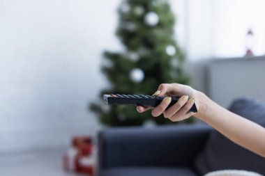 Uzaktan kumandalı, bulanık noel ağacının yanındaki kadın elinin kırpılmış görüntüsü.