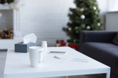 Kağıt peçeteli masa, hap kutusu, elektronik termometre ve bulanık noel ağacının yanındaki akıllı telefon.