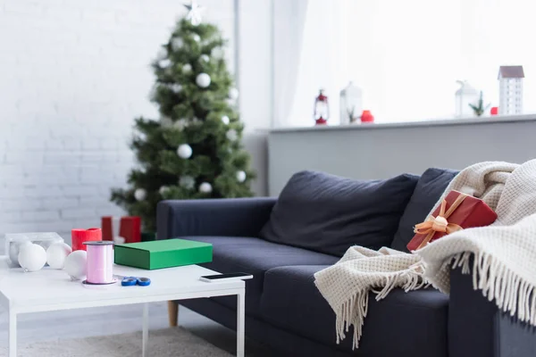 blanket and gift box on sofa near table with decorative ribbon and scissors in living room with blurred christmas tree