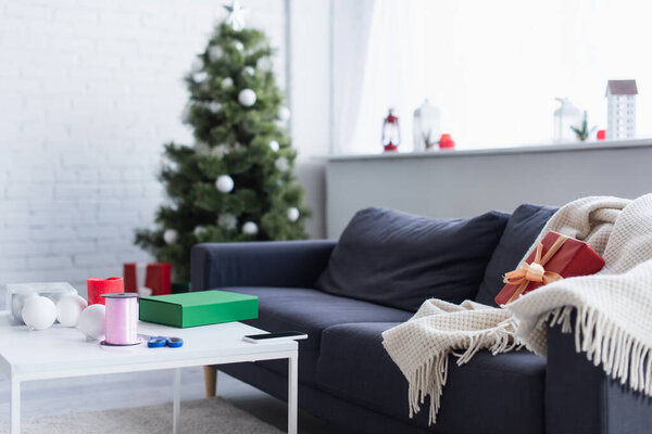blanket and gift box on sofa near table with decorative ribbon and scissors in living room with blurred christmas tree