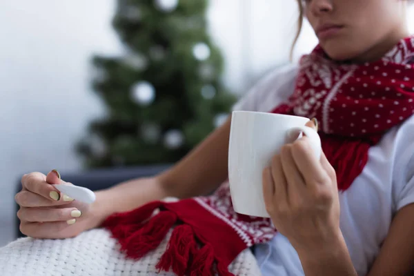 体温計のついた病気の女性と背景のぼやけたクリスマスツリーの近くの暖かい飲み物のカップのクロップドビュー — ストック写真