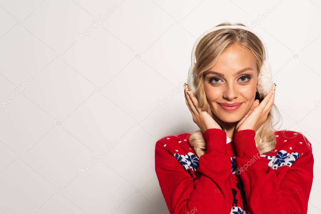 smiling blonde woman in red sweater touching earmuffs while looking at camera on white