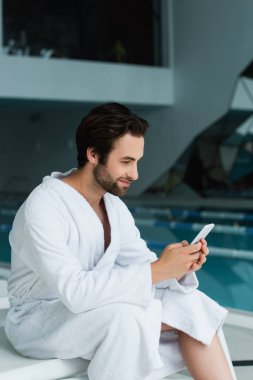 Bornozlu bir adam kaplıcadaki iskemlede cep telefonu kullanıyor. 