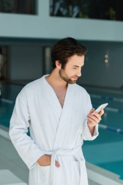 Bornozlu genç adam spa merkezinde yüzme havuzunun yanında akıllı telefon kullanıyor. 