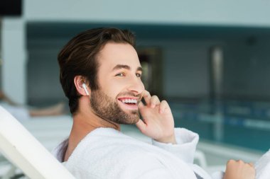 Kablosuz kulaklıklı ve bornozlu gülümseyen adam spa merkezinde iskemlede dinleniyor. 