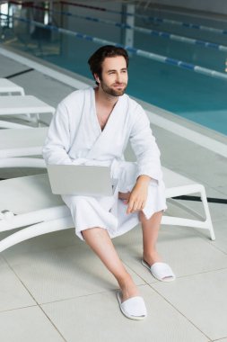 Bornozlu ve kulaklıklı bir adam spa merkezindeki iskemlede dizüstü bilgisayar tutuyor. 