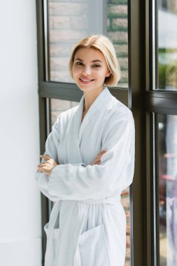 Smiling woman in bathrobe standing with crossed arms near window in spa center  clipart