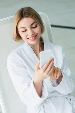 Bornozlu gülümseyen kadın spa merkezinde güvertedeki sandalyede akıllı telefon kullanıyor. 