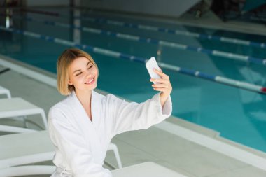 Gülümseyen kadın spa merkezinde yüzme havuzunun yanında akıllı telefondan selfie çekiyor. 