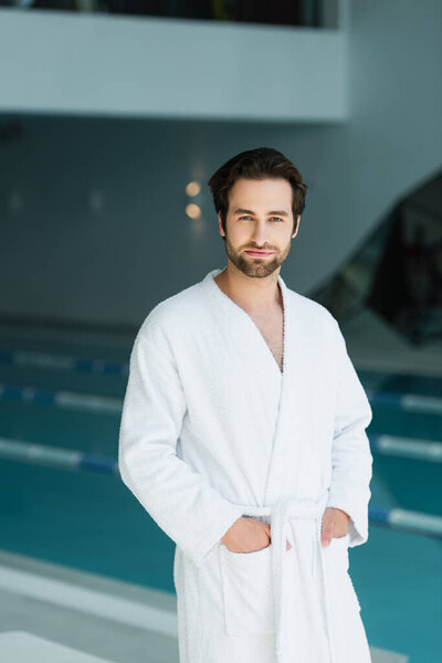 Young man holding hands in pockets of bathrobe in spa center 