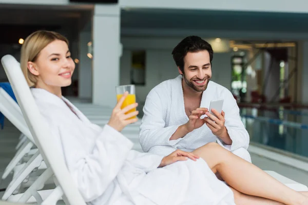 Smiling Man Bathrobe Using Cellphone Blurred Girlfriend Orange Juice Spa — Stock Photo, Image