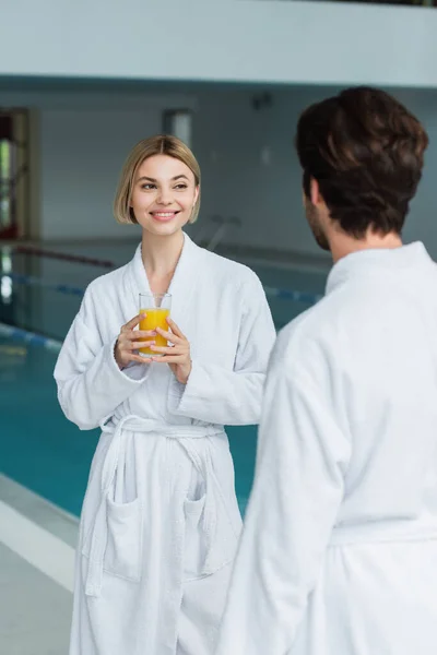 在温泉中心的白色浴衣中 一个快乐的女人在朦胧的男友身边拿着一杯橙汁 — 图库照片