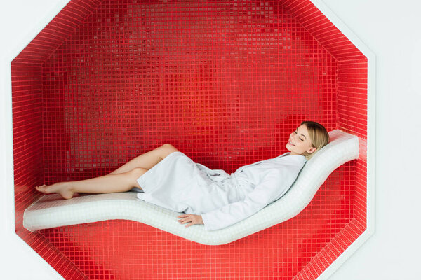 Cheerful woman in white bathrobe lying on lounge chair in spa center 