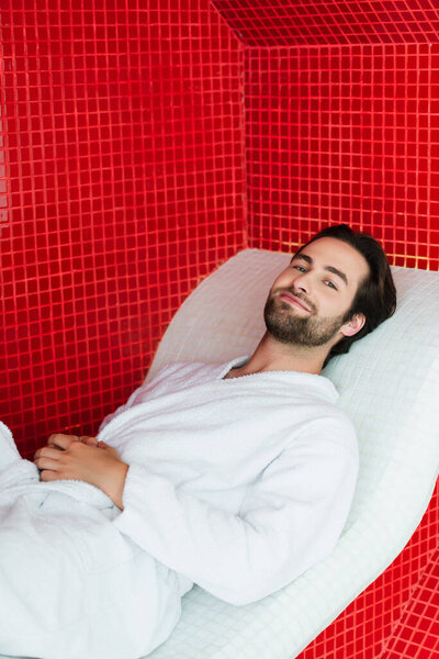 Young man in bathrobe relaxing on lounge chair in spa center 
