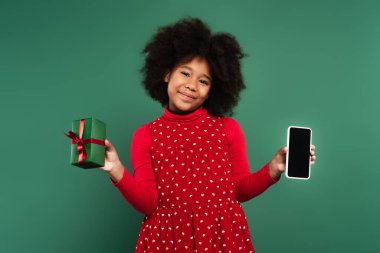 Kırmızı elbiseli, gülümseyen Afrikalı Amerikalı çocuk yeşil ekranda boş ekranlı akıllı telefon hediye ediyor. 