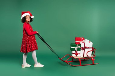 Noel Baba şapkalı afro-amerikan kızı yeşil arka planda hediyelerle kızak çekiyor. 