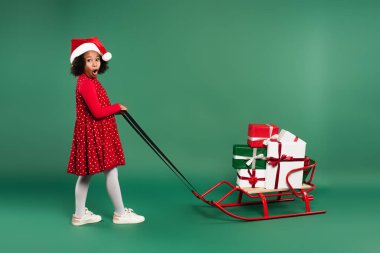 Amazed african american girl in santa hat pulling sleigh with gifts on green background  clipart