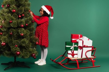 Gülümseyen Afrikalı Amerikalı kız, Noel Baba şapkasıyla Noel ağacını yeşil arka planda hediyelerle süslüyor. 