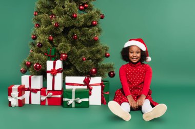 Noel Baba şapkalı neşeli Afro-Amerikalı çocuk yeşil arka plandaki Noel ağacının altında hediyelere bakıyor. 
