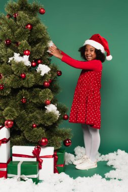 Noel Baba şapkalı ve kırmızı elbiseli Afrikalı Amerikalı kız Noel ağacının yanında dekoratif kar ve yeşil arka planda hediyeler tutuyor. 