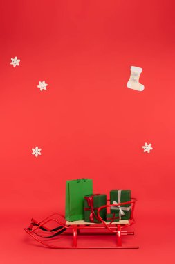 Gifts and shopping bag on sleigh near snowflakes on red background clipart
