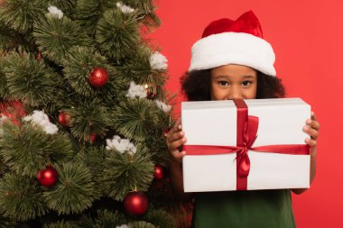 Gülümseyen Afrikalı Amerikalı çocuk Noel Baba şapkası takmış, elinde hediye tutuyor ve kırmızı üzerinde izole edilmiş noel ağacı var. 