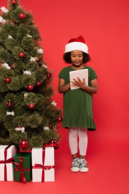 Noel Baba şapkalı gülümseyen Afrikalı Amerikalı çocuk hediye kutularının yanında dijital tablet ve kırmızı arka planda noel ağacı kullanıyor.