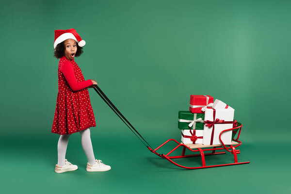 Amazed african american girl in santa hat pulling sleigh with gifts on green background 