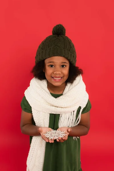 Stock image African american child in warm hat and scarf holding decorative snowflake and smiling isolated on red 