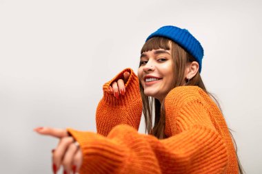 Samimi turuncu kazaklı ve mavi şapkalı genç kadın kamerayla neşe içinde poz veriyor ve etkileşime geçiyor..