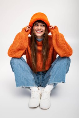 Güzel genç bir kadın rahat turuncu bir kazak giyer, neşeli bir pozda mutlu bir şekilde oturur..