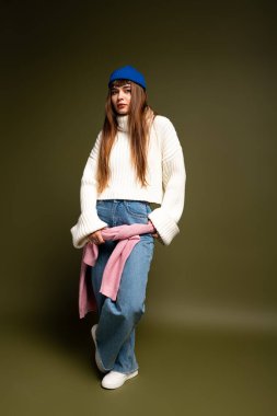 Güzel genç bir kadın, şık sonbahar kıyafetini kapalı bir ortamda gülümseyerek gösteriyor..