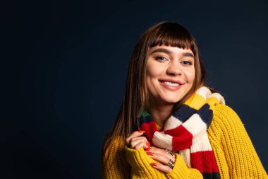 Samimi bir süveter ve atkı giymiş genç bir kadın koyu bir arka plana karşı mutlu bir şekilde ışıldıyor..