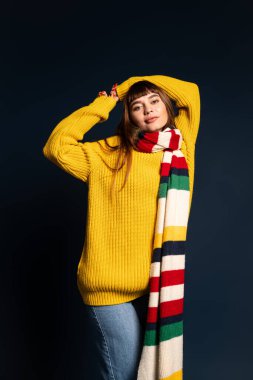 Canlı bir süveter ve eşarp giyen genç bir kadın, esmer bir arka plana şakayla poz veriyor..