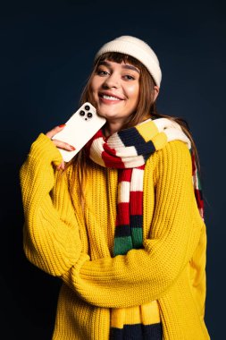 Hoş süveter ve eşarp giymiş güzel genç bir kadın telefonuyla poz verirken neşeli bir anı paylaşıyor..