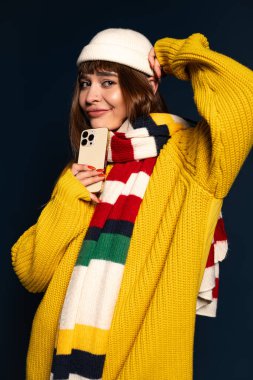 Parlak giyimli genç bir kadın telefonunu tutarken ve sıcak poz verirken neşeyle gülümsüyor..