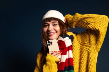 Sıcak giyimli genç bir kadın telefonuyla karanlık bir arkaplanda mutlu bir poz veriyor..