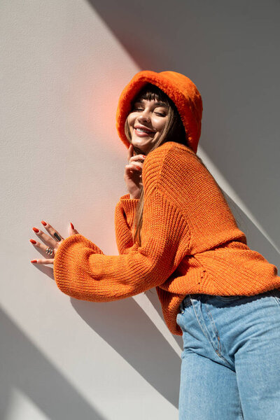 Радостная молодая женщина позирует с улыбкой, демонстрируя свой уютный осенний наряд в солнечном пространстве.