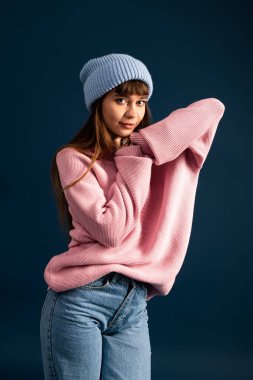 Pembe kazaklı ve mavi bereli genç bir kadın rahat bir yerde canlı poz veriyor..