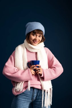 Sıcak ve şık giyinen genç kadın, sonbaharın sıcacık havasını kucaklayan mavi bir kupa tutuyor..