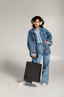 Chic woman poses with a shopping bag, flaunting her stylish denim outfit and headphones.