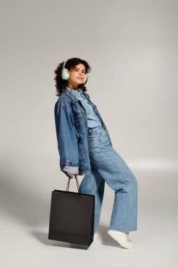 A young woman in denim poses confidently while carrying a shopping bag, embracing her fashion sense.
