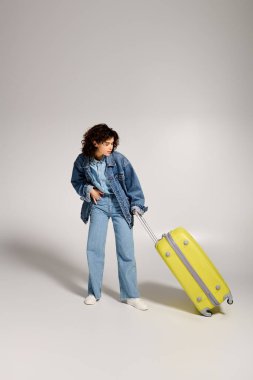 A beautiful young woman in a denim outfit stands with a bright yellow suitcase, ready for adventure.