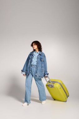 Young woman stands confidently with a bright suitcase, showcasing her trendy denim outfit.