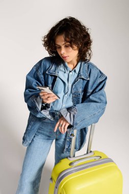 Stylish woman with curly hair checks her phone near a bright yellow suitcase, ready to travel.