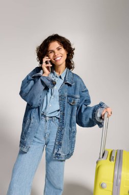Fashionable young woman smiles while chatting on her phone, holding a vibrant suitcase in hand.