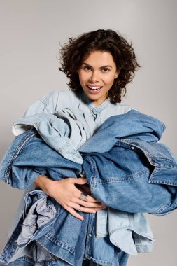 A young, beautiful woman joyfully holds a collection of denim jackets, showcasing her trendy style.